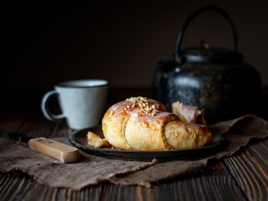 A glazed cardamom bun on a dark plate beside a mug and a cast iron teapot