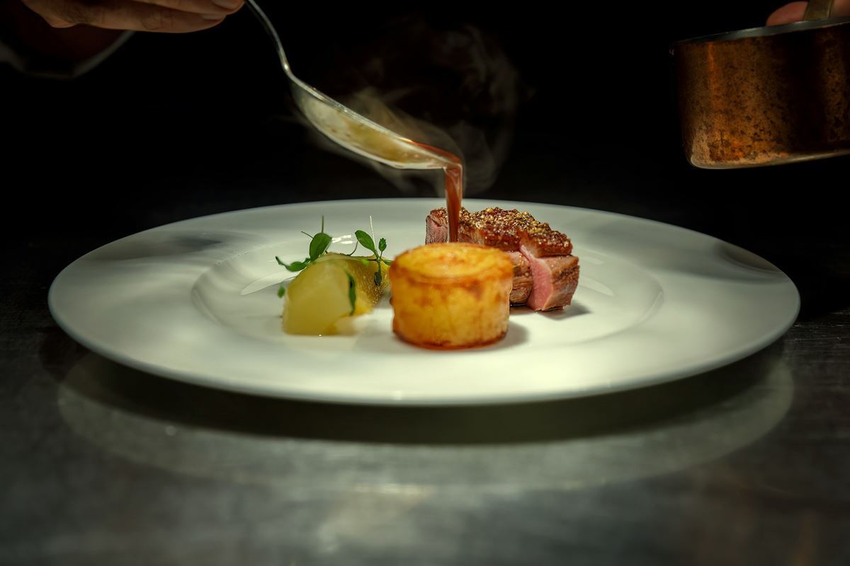A chef finishing a plated main course with sauce poured from a copper pan