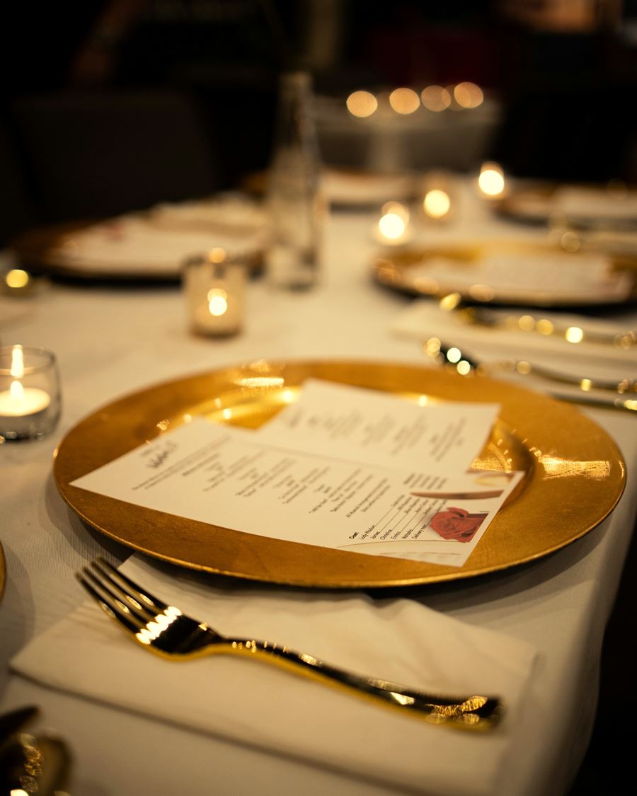 A printed evening menu resting on a brass charger, lit by candles on a linen tablecloth